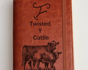 May include: A brown leather journal with a  'Twisted Y Cattle' inscription and a cow and calf illustration. The journal has a  'F' monogram and a brown elastic closure.
