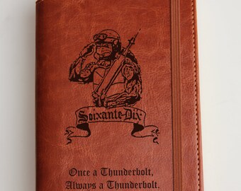 Pode incluir: Um diário de couro marrom com uma inscrição personalizada. A inscrição inclui um gráfico de um soldado, o texto "Soixante-Dix" e a citação "Once a Thunderbolt, Always a Thunderbolt. Brittany A. Anketell".
