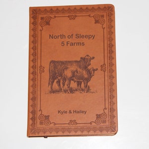 May include: A brown leather-bound journal with an engraved design. The cover features a decorative frame with the text "North of Sleepy 5 Farms" and an image of two cows. The bottom of the cover reads "Kyle & Hailey".