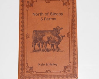 May include: A brown leather-bound journal with an engraved design. The cover features a decorative frame with the text "North of Sleepy 5 Farms" and an image of two cows. The bottom of the cover reads "Kyle & Hailey".