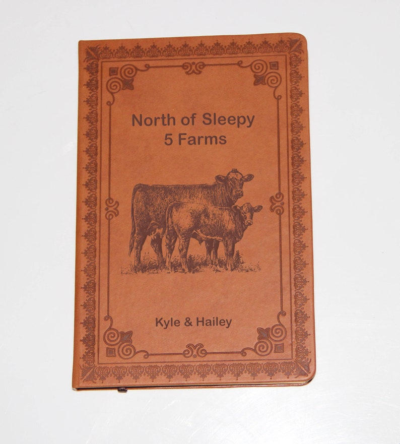 May include: A brown leather-bound journal with an engraved design. The cover features a decorative frame with the text "North of Sleepy 5 Farms" and an image of two cows. The bottom of the cover reads "Kyle & Hailey".