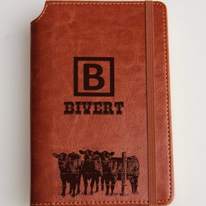 May include: Brown leather journal with a debossed design. The cover features a large letter "B" inside a square, the name "BIYERT", and a graphic of cows behind a fence. A brown elastic band secures the journal.