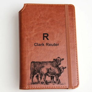 May include: Brown leather journal with the engraved text "R Clark Reuter" and an illustration of two cows. The journal has a brown elastic band closure.