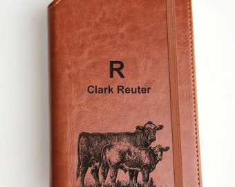 May include: Brown leather journal with the engraved text "R Clark Reuter" and an illustration of two cows. The journal has a brown elastic band closure.