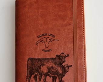 May include: A brown leather journal with a stitched border and a black elastic closure. The front cover features an engraved design of a cow and calf with the text "CALLAHAN CATTLE COMPANY" and a number "3" above the cow's head.