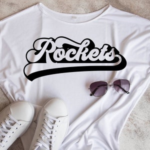 May include: White t-shirt with a black graphic that says "Rockets" in a retro font. The shirt is laying flat on a textured surface with a pair of sunglasses and white sneakers.