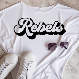 May include: White t-shirt with the word "Rebels" printed in black retro-style lettering. The shirt is laid flat on a light brown surface with a pair of sunglasses and white sneakers.