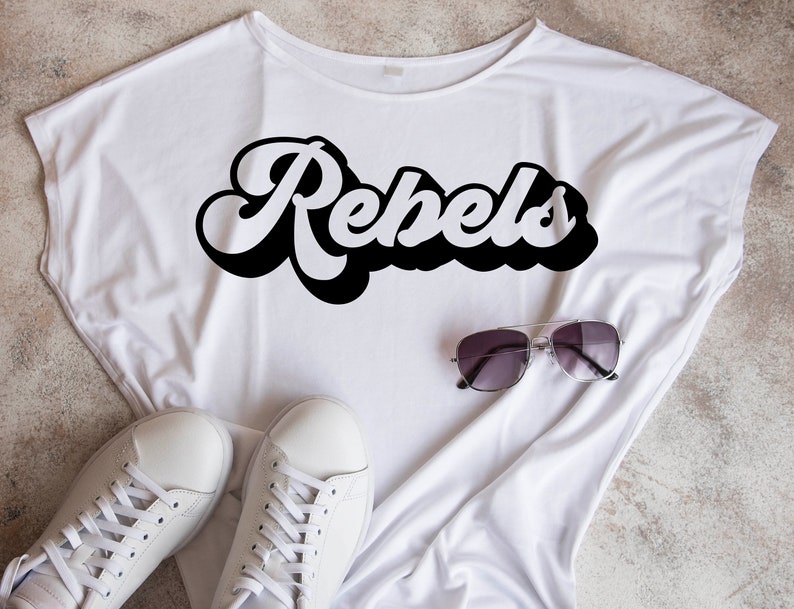 May include: White t-shirt with the word "Rebels" printed in black retro-style lettering. The shirt is laid flat on a light brown surface with a pair of sunglasses and white sneakers.