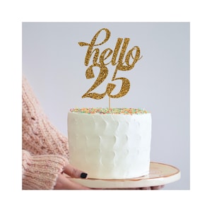 Può includere: Topper per torta con glitter dorati con il testo "hello 25" su una torta bianca.