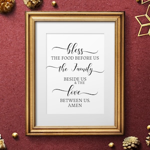 Può includere: Una stampa incorniciata con una cornice dorata e uno sfondo bianco. La stampa presenta un design di testo in bianco e nero che recita "bless the food before us the family beside us & the love between us. amen".
