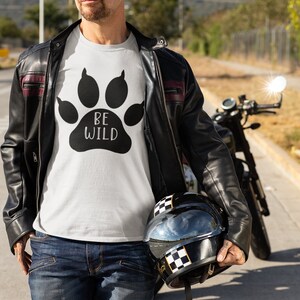 Può includere: Un uomo indossa una maglietta bianca con una grafica di un'impronta di zampa nera e il testo "BE WILD". Indossa anche una giacca di pelle nera e jeans blu. Sta tenendo un casco da motocicletta nero con una striscia a scacchi bianca.