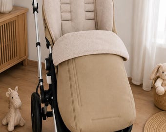 May include: A beige stroller featuring a soft, cream-colored fleece seat liner and a matching footmuff. The stroller has a black frame, a brown leather-look handlebar, and four black wheels. The seat is upright with integrated harness slots.
