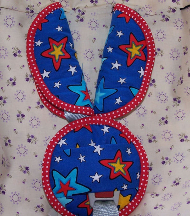 May include: A set of blue fabric harness covers featuring a pattern of red, yellow, and light blue stars with white accents. The covers have a rounded shape with red and white polka dot trim along the edges and include a central buckle component.