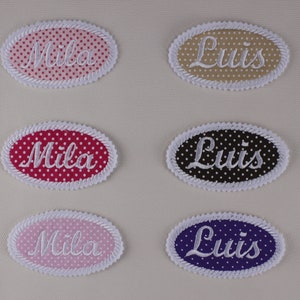 Peut inclure: Six patchs en tissu ovales avec des pois blancs et des noms brodés en blanc. Les noms sont "Mila" et "Luis". Les patchs sont en rose, beige, rouge, noir, rose clair et violet.