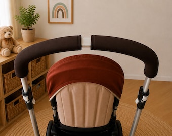 May include: A stroller handlebar cover set in dark brown, featuring two separate foam-like sleeves designed to fit over the curved metal frame of a stroller handle. The covers provide a soft, matte texture and a secure grip for the stroller.