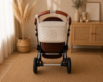 May include: A cream-colored quilted diaper bag with a brown interior is attached to a stroller. The bag has a circular monogram with the letters "JS". The stroller has brown leather handles and a brown seat. The wheels are black.