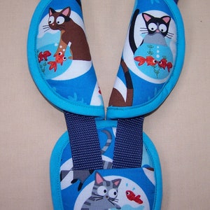 May include: A set of blue fabric harness strap covers featuring a repeating pattern of cartoon cats and goldfish in bowls. The covers have bright blue trim and are attached to dark navy blue nylon webbing straps, designed for use on a harness system.