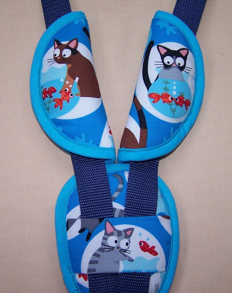 May include: A set of blue fabric harness strap covers featuring a repeating pattern of cartoon cats and goldfish in bowls. The covers have bright blue trim and are attached to dark navy blue nylon webbing straps, designed for use on a harness system.
