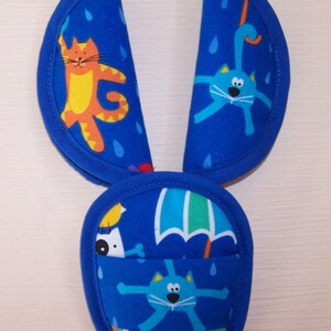 May include: A blue fabric seat belt cover set featuring a repeating pattern of colorful cartoon cats, raindrops, and umbrellas. The set includes two elongated rectangular pads with rounded ends and a central rounded pouch with blue trim.