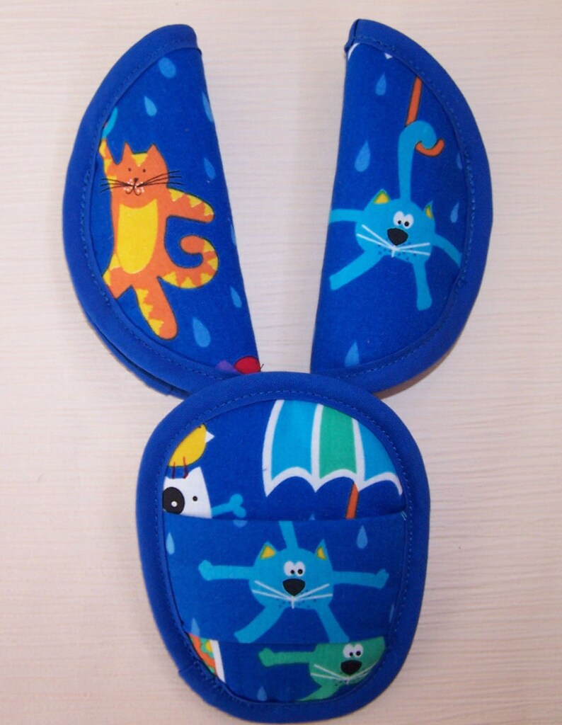 May include: A blue fabric seat belt cover set featuring a repeating pattern of colorful cartoon cats, raindrops, and umbrellas. The set includes two elongated rectangular pads with rounded ends and a central rounded pouch with blue trim.
