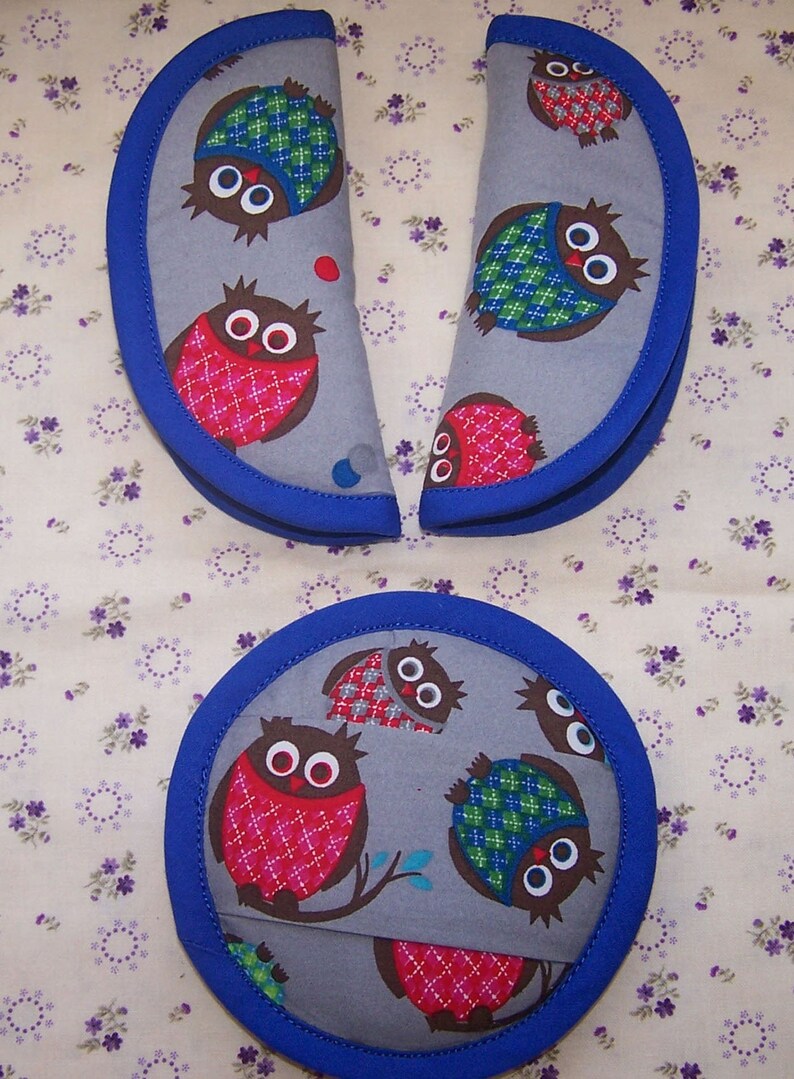May include: Two semi-circular fabric pads and one circular fabric pad, all featuring a gray background with a repeating pattern of brown owls wearing red or green plaid vests, finished with a solid blue fabric trim along the curved edges.