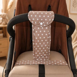 May include: A beige pushchair crotch strap cover featuring a white star pattern. The rectangular fabric cover wraps around the centre harness strap and includes a rounded top flap secured with a single white snap button closure.