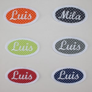 Puede incluir: Parches bordados de forma ovalada en varios colores, incluyendo naranja, azul marino, rojo, verde y gris. Cada parche presenta un borde blanco con forma de cuerda y un fondo de lunares. Los parches muestran nombres como "Luis" y "Mila" en escritura blanca.