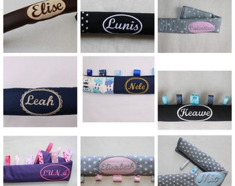 May include: A collection of personalized baby accessories, including stroller straps and sensory ribbons. The straps are in various colors, such as navy, black, and gray, and feature embroidered names like "Elise," "Lunis," and "Leah." The sensory ribbons are in various colors and patterns.