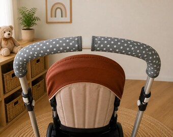 Könnte beinhalten: Ein Kinderwagen-Lenkerbezug-Set aus grauem Stoff mit weißem Sternenmuster. Die Bezüge sind zylindrisch und umschließen die beiden gebogenen Metallgriffe des Kinderwagens, der vor einem rostfarbenen und beigen Sitzverdeck positioniert ist.