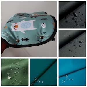 Könnte beinhalten: Ein Paar grüne Kinderwagen-Handwärmer mit einem weißen Bären- und Waldmuster. Die Handwärmer sind wasserdicht und haben einen schwarzen Besatz. Das Bild zeigt Nahaufnahmen des Stoffes mit Wassertropfen darauf.