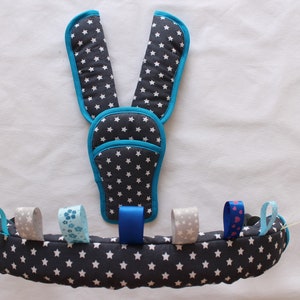 May include: A grey and white star patterned baby activity toy with blue trim and colourful ribbons. The toy has two shoulder pads and a circular base.