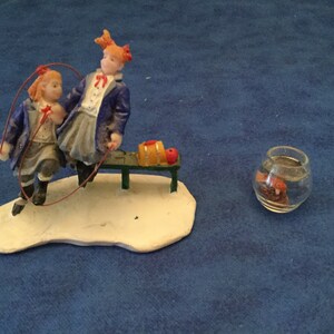 May include: A miniature figurine set depicting two girls in blue and white school uniforms, one jumping rope while the other sits on a bench with a red apple and a yellow book. A small glass bowl with a goldfish is in the background.