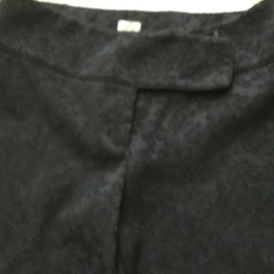 Puede incluir: Pantal&oacute;n negro de tela texturizada ligeramente brillante. El pantal&oacute;n tiene una cintura con bot&oacute;n y cierre de cremallera.