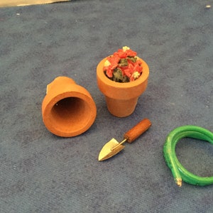 May include: A miniature gardening set with a terracotta pot, a small shovel, and a green hose. The pot is filled with red and white flowers.