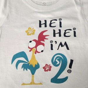 May include: White t-shirt with a cartoon rooster and the text "Hei Hei I'm 2!" printed on it. The rooster is red, yellow, and blue. The number 2 is blue and has a wave design. There are two yellow flowers on the shirt.