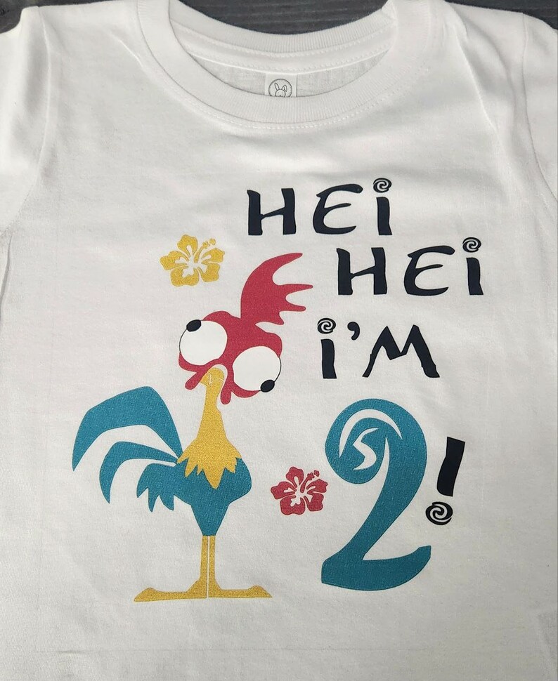 May include: White t-shirt with a cartoon rooster and the text "Hei Hei I'm 2!" printed on it. The rooster is red, yellow, and blue. The number 2 is blue and has a wave design. There are two yellow flowers on the shirt.