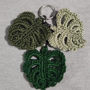 Puede incluir: Un llavero con tres hojas de monstera de ganchillo en tonos verdes. Las hojas están unidas a un llavero plateado.