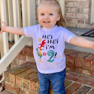 May include: A white t-shirt with a cartoon rooster and the text "Hei Hei I'm 2!" printed on it. The shirt is being worn by a young child.