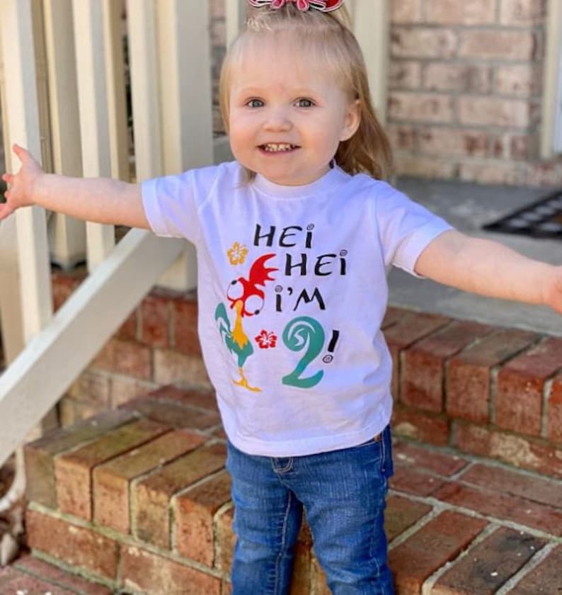May include: A white t-shirt with a cartoon rooster and the text "Hei Hei I'm 2!" printed on it. The shirt is being worn by a young child.