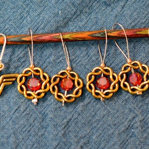 May include: Five gold-colored stitch markers with red glass beads in the center, and one gold-colored Wonder Woman charm. The stitch markers are hanging on a wooden knitting needle.