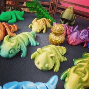 May include: A collection of colorful, 3D printed figurines, including a green lizard, a gold-painted bird, a purple dragon, and a green cat. The figurines are arranged on a black surface.