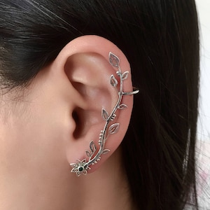 Op de afbeelding: Zilveren oor manchet met een gedetailleerd wijnstokontwerp, compleet met bladeren en een kleine bloem met een groen centrum. De manchet buigt rond het oor en biedt een elegant, door de natuur geïnspireerd accessoire.