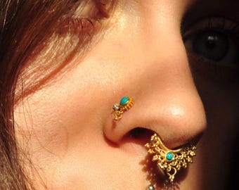 May include: A gold-toned nose ring featuring an intricate, fan-shaped filigree design adorned with a small, round turquoise stone. A matching gold-toned nostril stud with a turquoise stone is also visible, positioned above the septum ring.