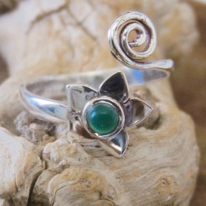 Può includere: Un anello d'argento con una pietra preziosa verde incastonata in un design floreale. L'anello è regolabile e presenta un dettaglio di filo attorcigliato.