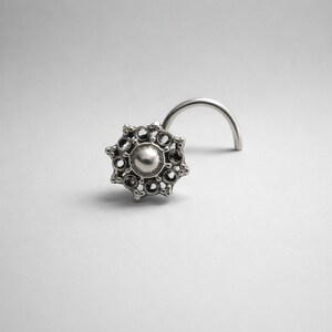 Pode incluir: Um piercing de nariz prateado com um design decorativo em forma de raio de sol. O piercing apresenta uma conta central redonda rodeada por pequenos detalhes circulares. A haste curva &eacute; vis&iacute;vel, projetada para perfura&ccedil;&atilde;o.