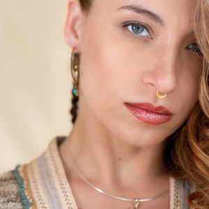 Puede incluir: Una mujer con ojos azules y cabello largo y castaño lleva un aro de nariz dorado y un par de pendientes dorados. Lleva un suéter de color beige y verde con estampado.