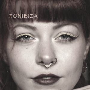 Op de afbeelding: Close-up van een vrouwengezicht met een septum neusring. De ring is goudkleurig en heeft een decoratief ontwerp. De afbeelding bevat ook het woord "RONIBIZA" bovenaan. De vrouw heeft donkerrode lippenstift en oogmake-up.