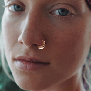 Puede incluir: Un aro nasal de tono dorado con un diseño semicircular y pequeñas protuberancias en forma de pétalo a lo largo del borde exterior. La joya está colocada en la fosa nasal, mostrando un acabado metálico y una estructura segmentada.