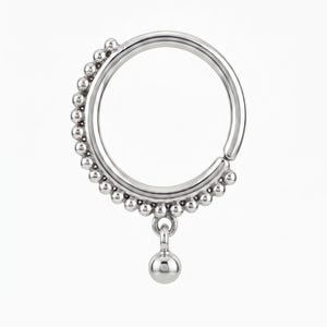 May include: A silver-coloured circular hoop ring featuring a row of small metallic beads along the outer edge. A single larger spherical bead hangs from the bottom of the hoop, suspended by a small jump ring attached to the beaded segment.