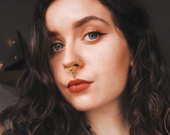May include: A close-up portrait of a person with wavy dark brown hair and blue eyes. The person is wearing gold-colored nose and lip piercings. The lips are painted with red lipstick. The person is also wearing a necklace.